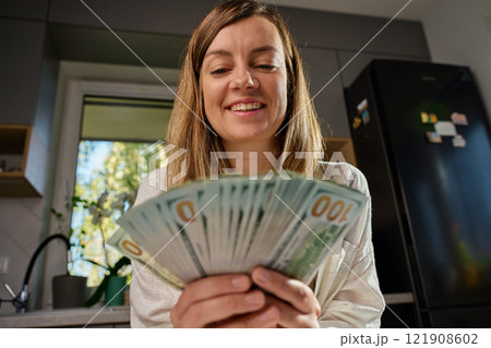 Smiling woman in kitchen counting cash while sitting at table with receipts and documents, managing home finances. Concept of planning home budget 121908602