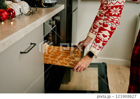Woman in red festive sweater putting tray of Christmas cookies into oven. Process of cooking traditional gingerbread cookies for winter holidays at home kitchen 121908627