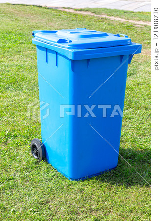Covered blue plastic garbage container with wheels on the green grass 121908710