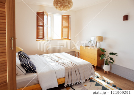 Sunny cozy bedroom with white walls. Wooden bed and chest of drawers with household things. A soft fluffy carpet and a potted houseplant. 121909291