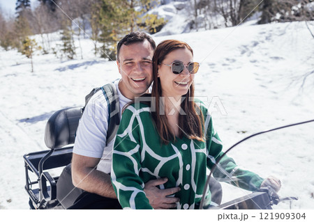 A young married couple is spending a vacation in the mountains. An attractive man and a woman are riding an ATV on a snowy road. 121909304