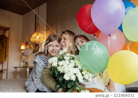 Laughing mom holds bouquet of flowers and colorful balloons, hugs her daughters. Young happy woman celebrates her birthday at home with her family. Laughing mom holds bouquet of flowers and colorful balloons, hugs her daughters. Young happy woman celebrates her birthday at home with her family. 121909331