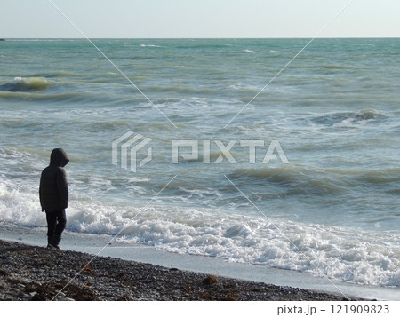 Child in jacket and boots walking along rocky shore of cold sea with grey-blue waves on cold winter or autumn day, lonely boy seen from back looking at surf 121909823