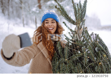 A young woman with a phone and a Christmas tree with garlands enjoys the beautiful winter weather. A young woman with a phone and a Christmas tree with garlands enjoys the beautiful winter weather. 121910750