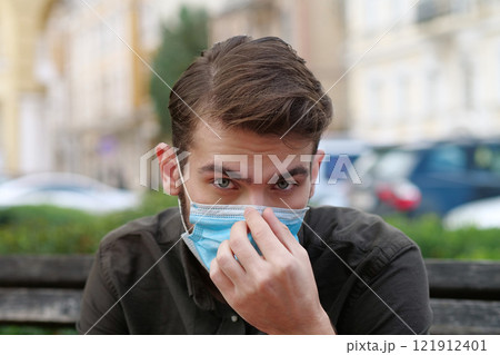 Young man sits on the bench in park and puts on medical protective face mask 121912401