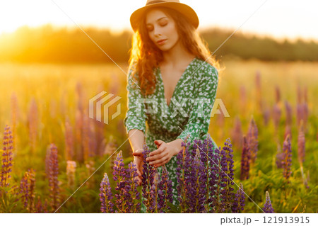 Close-up of young woman in hat and curly hair touching lupins in field at sunset. Fashion and nature Close-up of young woman in hat and curly hair touching lupins in field at sunset. Fashion and nature 121913915