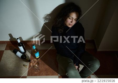 Woman sitting on tiled floor surrounded by wine bottles and a glass 121915016