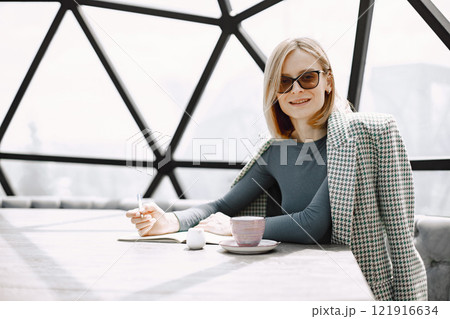 Indoor portrait of a young businesswoman sitting in a cafe and writing a documents. Blonde girl wearing sunglasses and jacket. There are glass wall behind. 121916634