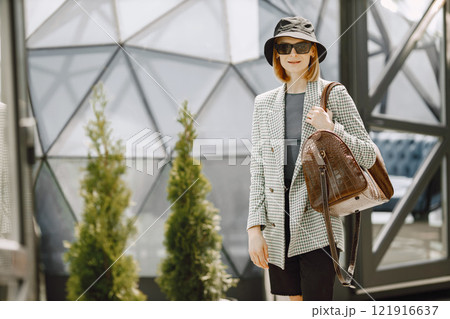 Outdoor portrait of a young beautiful fashionable woman walking on the street, holding big brown leather bag. Blonde girl wearing sunglasses and black hat. There are round glass building behind. 121916637