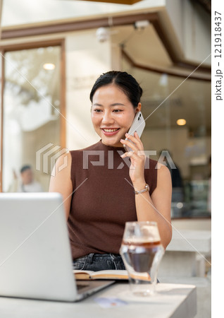 A beautiful, confident Asian woman talks in the phone while working remotely from a cafe. A beautiful, confident Asian woman talks in the phone while working remotely from a cafe. 121918437