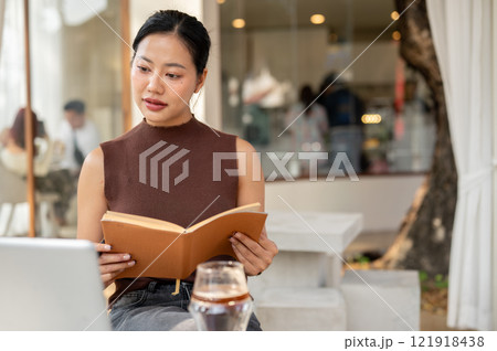 A positive Asian woman works remotely from a city cafe, researching information in a book. 121918438