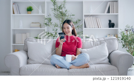 Attractive smiling young Asian woman using mobile phone while sitting on the sofa at home Attractive smiling young Asian woman using mobile phone while sitting on the sofa at home 121919140