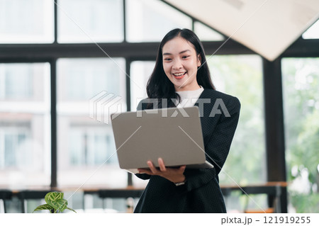 Confident Businesswoman Smiling While Holding Laptop in Modern Office with Large Windows and Natural Light Confident Businesswoman Smiling While Holding Laptop in Modern Office with Large Windows and Natural Light 121919255