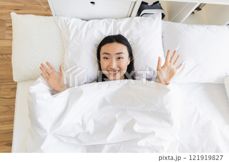 Smiling Asian woman in bed showing happiness and relaxation. Indoor morning scene with young woman, bright bedding, and cheerful expression, Asian female greeting new day with joy and positivity. Smiling Asian woman in bed showing happiness and relaxation. Indoor morning scene with young woman, bright bedding, and cheerful expression, Asian female greeting new day with joy and positivity. 121919827