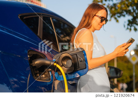 Woman in a white dress stands beside an electric car, checking her smartphone while it charges. Woman in a white dress stands beside an electric car, checking her smartphone while it charges. 121920674