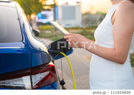 Pregnant woman in white dress charging an electric car and using her mobile phone to pay 121920686