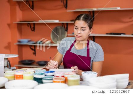 Woman potter with paintbrush glazing, painting on plate in workshop, working in pottery 121922667