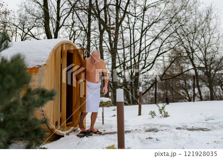 Older man comming out of small snow-covered sauna. Winter Spa weekend getaway for elderly people. 121928035