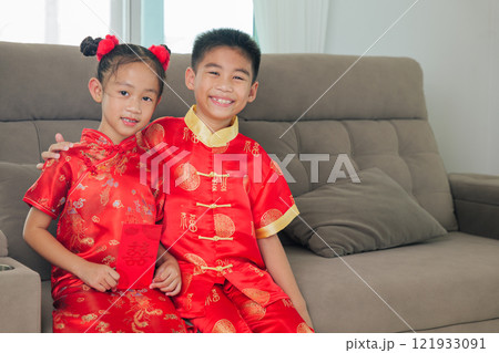 Smiling brother and sister in festive red outfits, holding red envelopes. They sit on a neutral-toned couch. A celebratory scene. Smiling brother and sister in festive red outfits, holding red envelopes. They sit on a neutral-toned couch. A celebratory scene. 121933091