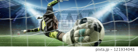 Goalkeeper in bright uniform extending his arm to catch soccer ball inches from net, with players and stadium visible in background under bright lights. Goalkeeper in bright uniform extending his arm to catch soccer ball inches from net, with players and stadium visible in background under bright lights. 121934048