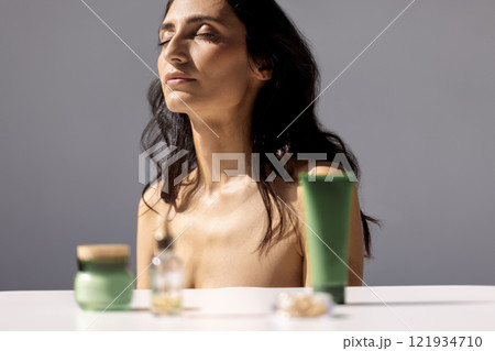 Close-up studio portrait of attractive young woman with her eyes closed in sunlight. Jars of cream and serum in foreground. Care about clean and soft face skin. 121934710
