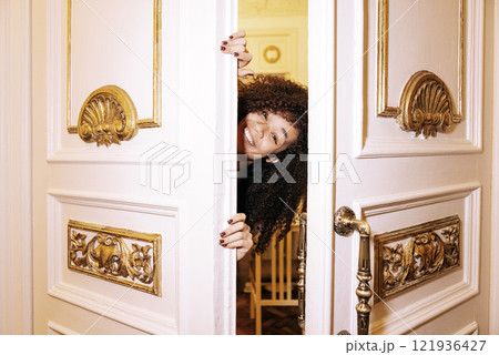 Young African in white elegant dress looks out from behind door. Joyful black girl laughs and greets guests in doorway. Teenager is having fun and waiting for friends. 121936427