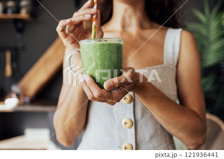 A transparent cup with a green smoothie in the hands of a young woman. A girl in a linen sundress drinks a fresh drink made from herbs and vegetables in the kitchen. A transparent cup with a green smoothie in the hands of a young woman. A girl in a linen sundress drinks a fresh drink made from herbs and vegetables in the kitchen. 121936441