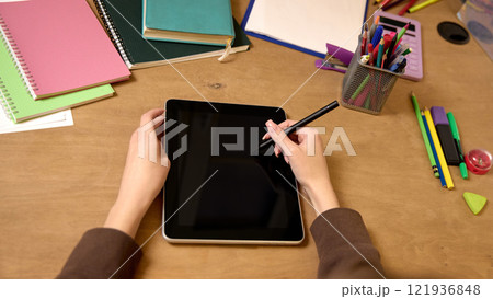 Overhead view of a woman using a stylus on a tablet at a desk with notebooks, stationery, and a calculator, emphasizing technology and education. 121936848