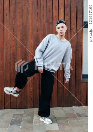 A young man wearing a gray sweatshirt and black pants stands in front of a wooden wall, his right leg raised in a relaxed pose A young man wearing a gray sweatshirt and black pants stands in front of a wooden wall, his right leg raised in a relaxed pose 121937169