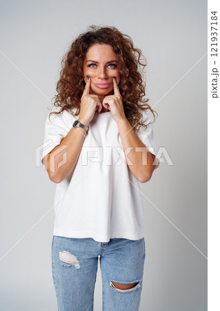 Cheerful woman playfully making a silly face while posing in casual attire against a plain backdrop Cheerful woman playfully making a silly face while posing in casual attire against a plain backdrop 121937184