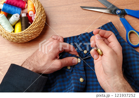 A person sewing a button on a blue plaid shirt with sewing tools and threads on a wooden table 121937252