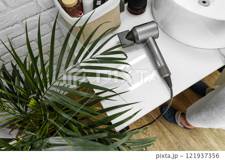 A modern hairdryer placed on a bathroom countertop surrounded by beauty products and a green plant A modern hairdryer placed on a bathroom countertop surrounded by beauty products and a green plant 121937356