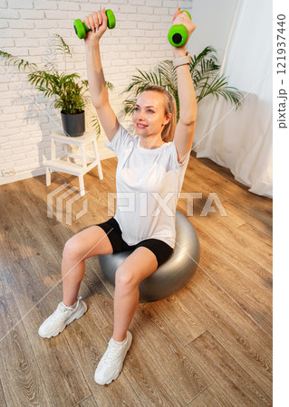 Woman exercising with dumbbells on a stability ball in a indoor space Woman exercising with dumbbells on a stability ball in a indoor space 121937440