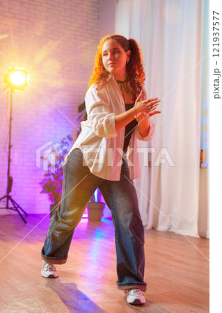 A young woman with curly red hair dances in a studio, wearing a white shirt and blue jeans A young woman with curly red hair dances in a studio, wearing a white shirt and blue jeans 121937577