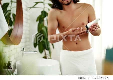 A young man applying skincare cream on his face with a focused expression, demonstrating personal grooming at home 121937971