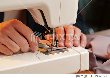 Hands sewing fabric using a sewing machine on a wooden table in a well-lit workspace 121937975