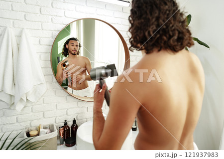 A man with curly hair styles himself in a bright room while using a hairdryer to enhance his look 121937983