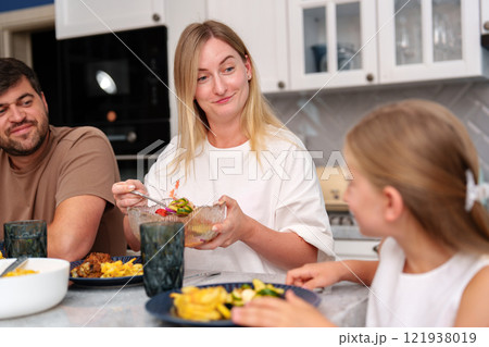 A woman sits at a kitchen table with her family, enjoying a meal together 121938019