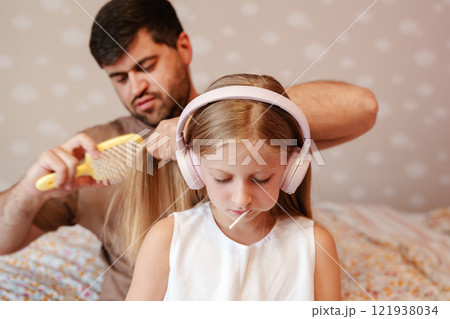 Father combing daughters long hair while she enjoys music with headphones 121938034