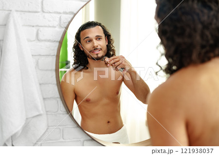 A man with curly hair grooming himself in the bathroom while looking into a round mirror 121938037