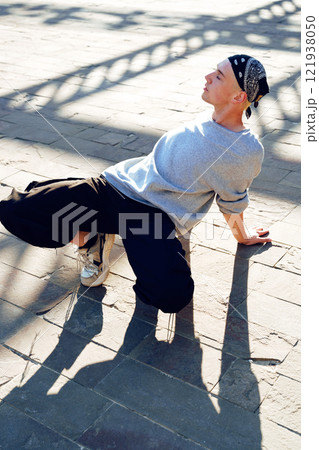 A young man practices a dance move on a sunny day, his long shadow stretching across the paved ground 121938050