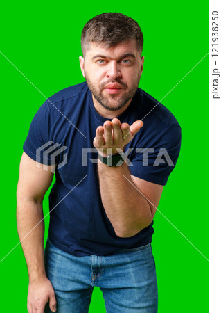 A man blowing a kiss while standing against a bright green backdrop in a casual outfit 121938250