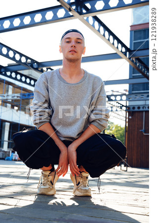 A man wearing a grey sweatshirt, black pants, and white sneakers is squatting on a paved surface A man wearing a grey sweatshirt, black pants, and white sneakers is squatting on a paved surface 121938289
