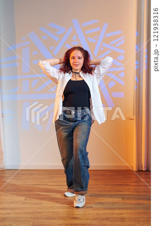 A young woman with curly red hair poses in a white shirt, black tank top, and blue jeans in front of a light projection 121938316
