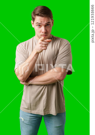 A young man with a serious expression pointing towards the viewer, standing against a bright green background A young man with a serious expression pointing towards the viewer, standing against a bright green background 121938666