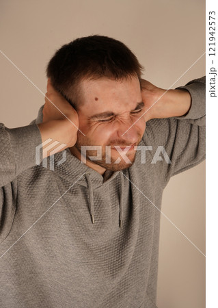 Man in depression. Portrait of young guy closing his ears by hands and keeping his eyes closed while isolated on grey. Photo of desperate annoyed bearded man keeps hands on head and closes eyes. Man in depression. Portrait of young guy closing his ears by hands and keeping his eyes closed while isolated on grey. Photo of desperate annoyed bearded man keeps hands on head and closes eyes. 121942573