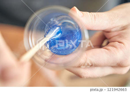 Close up of hands mixing blue epoxy resin in clear plastic cup with wooden 121942663