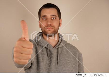 Charming confident guy dressed gray hoodie showing thumb up alone. Young handsome man with beard over isolated gray background giving a thumbs up gesture. Okay sign. Agree. 121942820