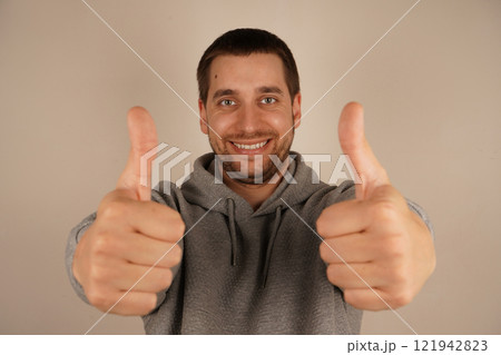 Young handsome man with beard over isolated gray background giving a thumbs up gesture. Approving expression looking at the camera. Charming confident guy dressed gray hoodie showing thumbs up alone. Young handsome man with beard over isolated gray background giving a thumbs up gesture. Approving expression looking at the camera. Charming confident guy dressed gray hoodie showing thumbs up alone. 121942823