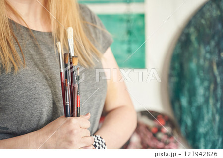 Caucasian female artist holding paintbrushes in studio with artworks Caucasian female artist holding paintbrushes in studio with artworks 121942826